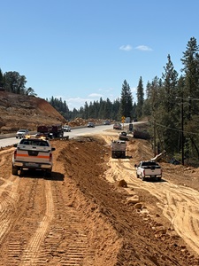 A highway construction zone with several work trucks and equipment positioned along a wide dirt embankment beside the road. Vehicles travel on the paved highway to the left, while pickup trucks and a dump truck move along the rough dirt access path to the right. Large piles of earth and rocks line the construction area, and tall pine trees stand in the background under a clear blue sky.