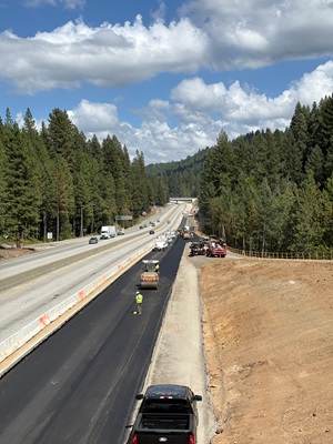 A road construction scene showing freshly paved black asphalt in a new lane beside an active highway. Several workers and paving machines operate along the new surface, while vehicles drive on the existing highway to the left. The area is surrounded by dense pine forest and rolling hills under a bright sky with large, scattered clouds.