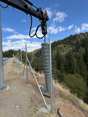 A roll of metal wire fencing is being lifted upright by a hydraulic arm attached to heavy equipment. The fencing is positioned beside a series of metal fence posts along the edge of a steep hillside covered in dense green trees. The sky above is bright blue with scattered white clouds.