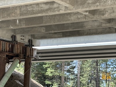 A close-up view beneath a concrete bridge showing support beams, metal brackets, and temporary construction shoring. Rust-colored steel posts and a green bracing pipe hold up a section of the structure. Pine trees are visible in the background through the gaps under the bridge.