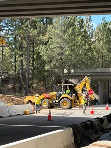 A construction worker holds a handheld stop sign while standing near a yellow backhoe loader digging beside concrete barriers under an overpass. Another worker in a high‑visibility vest stands near the equipment, and orange traffic cones line the work area. Tall pine trees fill the background.