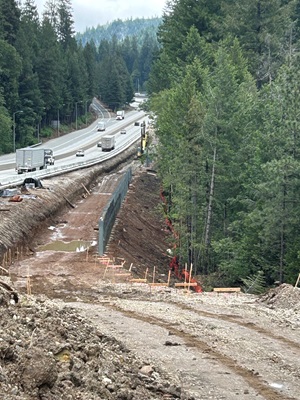 A construction work zone runs alongside a forested highway in a mountainous area. Heavy equipment is grading a long, narrow embankment next to the roadway, with freshly excavated dirt, erosion‑control fencing, and survey stakes visible. Traffic, including trucks and passenger vehicles, travels on the adjacent multi‑lane highway. Tall pine trees and rolling hills surround the site under an overcast sky.