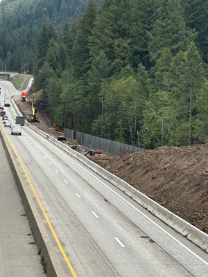 A view of a highway construction zone running alongside a forested mountainside. Traffic moves on the open lanes while heavy equipment works on an excavated embankment separated by concrete barriers. Freshly graded soil, erosion‑control fencing, and construction materials line the work area, with dense evergreen trees rising on the hillside beside the roadway.