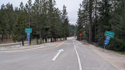 A two‑lane roadway leads toward a freeway on‑ramp surrounded by tall pine trees. Green signs on both sides of the road indicate the entrance to Interstate 80 East. A white arrow is painted on the pavement, guiding traffic forward. In the distance, an orange construction warning sign is visible near the start of the on‑ramp.