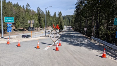 A highway on‑ramp is blocked by orange traffic cones and a “Ramp Closed” sign. The closure is set on a paved ramp surrounded by tall pine trees. Additional road signs indicate the entrance to Interstate 80 East, and vehicles can be seen traveling on the adjacent freeway below. The scene is outdoors on a clear day with blue sky.
