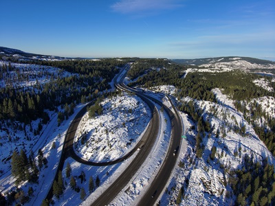 A high-angle aerial view of a snow-covered mountain landscape with a highway interchange running through the scene. The roads curve between forested slopes, with patches of snow on the ground and trees. A few buildings and vehicles are visible near the interchange. In the distance, rolling mountains stretch beneath a cloudy sky with soft light breaking through.