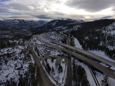A high-angle aerial view of a snow-covered mountain landscape with a highway interchange running through the scene. The roads curve between forested slopes, with patches of snow on the ground and trees. A few buildings and vehicles are visible near the interchange. In the distance, rolling mountains stretch beneath a cloudy sky with soft light breaking through.