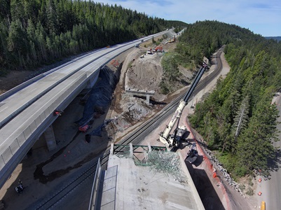 A wide aerial view of a highway bridge demolition and reconstruction site in a forested mountain area. One intact elevated roadway runs along the left side, while a partially demolished bridge deck appears in the foreground with exposed rebar. Below, active railroad tracks run through the work zone. A large crane and multiple construction vehicles are positioned on the right side, where crews are clearing earth and preparing new bridge supports. Dense evergreen trees cover the surrounding hills under a bright sky.