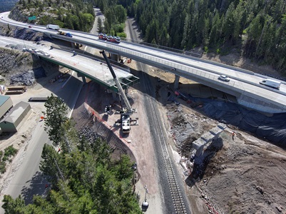 A high‑angle view of an active bridge demolition and reconstruction site in a forested mountain corridor. A partially dismantled bridge spans over a roadway and active railroad tracks, with exposed steel beams and construction barriers visible. A large crane is positioned below the bridge, lifting materials near the tracks. Adjacent to the demolished span, traffic moves across a newer parallel overpass. Construction vehicles and equipment are staged around the site, and dense pine trees cover the surrounding hillsides.