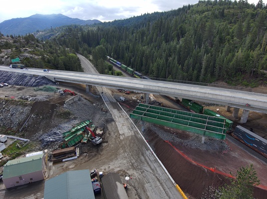 A high‑angle aerial view of a bridge demolition and reconstruction site in a mountainous, forested area. A completed highway overpass carries traffic above the work zone, while a partially dismantled green steel bridge structure lies below. Construction equipment and debris are spread across the site, including excavators and piles of removed steel beams. Multiple freight train cars travel along the railroad tracks that run through the center of the scene. Tall pine trees and rugged terrain surround the active construction area under a cloudy sky.