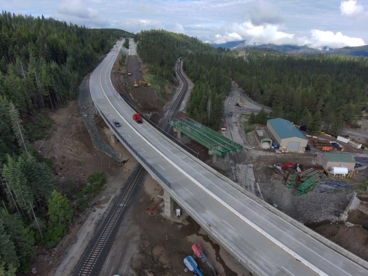 A high‑angle aerial view of a highway bridge construction site in a mountainous, forested area. A newly built elevated roadway curves through the scene with vehicles traveling on it. Below, railroad tracks run parallel to the structure. Nearby, construction equipment, dirt work, and a partially dismantled green steel bridge span are visible. Dense pine forests cover the surrounding hills, and clouds hang over distant mountain peaks.