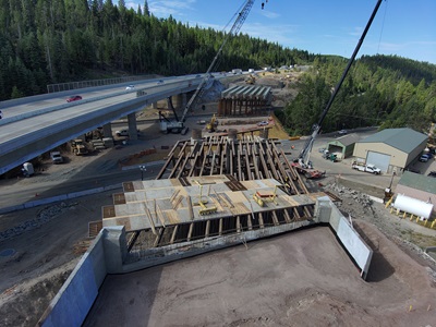 An overhead view of a highway construction site in a forested mountain area. Crews and heavy equipment, including cranes and excavators, are working on a new bridge structure beneath an existing overpass. Wooden and steel framework is being assembled, and concrete retaining walls are visible in the foreground. Traffic continues on the elevated roadway while construction progresses alongside.