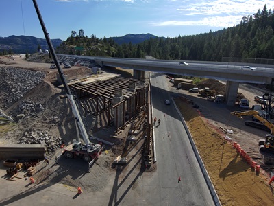 A highway construction site beneath an overpass in a forested mountain area. Crews are working around large equipment, including a crane and excavator, as new bridge support structures are being built beside the roadway. Traffic is limited to one direction while workers and machinery operate near the partially completed bridge.