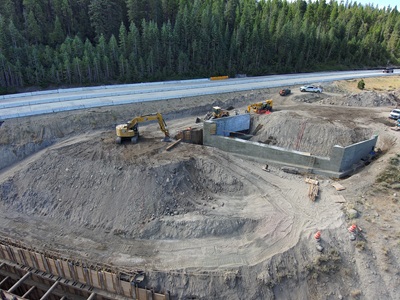 An aerial view of a highway construction site surrounded by dense forest. Heavy machinery, including excavators and loaders, are working around newly built concrete retaining walls and large dirt embankments. Graded earth and tire tracks cover the work area, with construction materials and vehicles positioned nearby. A completed section of highway runs along the top edge of the image.