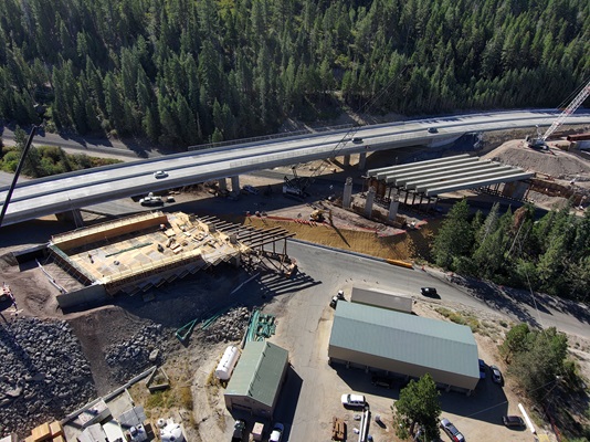 An aerial view of a bridge construction site in a forested mountain area. A completed highway bridge with cars traveling on it runs across the top of the image. Below it, a new bridge structure is being built, with large concrete columns, steel girders, and construction equipment including cranes and excavators. One section shows wooden framing and decking under construction. Several small buildings, parked vehicles, and materials are visible in the lower portion of the image. Tall pine trees surround the entire area.