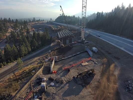 An aerial view of a highway construction site in a mountainous forested area. A large crane stands near partially built bridge supports, with workers and construction equipment spread around the site. A nearby roadway curves along the hillside, and railroad tracks run below the construction zone. Early morning light and haze create soft shadows across the landscape.
