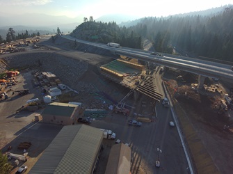 Aerial view of a highway bridge construction site in a forested mountain area. A new bridge deck is being formed with wooden framing and green rebar beneath an elevated roadway carrying traffic. Construction materials and equipment are staged around the site, with rocky slopes and tall pine trees in the background.