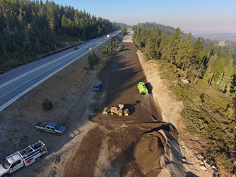 Aerial view of a highway construction area beside a forested mountainside. Heavy equipment and trucks work on grading a wide dirt section parallel to the roadway, while vehicles travel on the adjacent highway. Trees line the slope on the right under clear morning light.