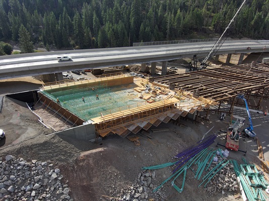 Aerial view of a bridge construction site in a forested area. Workers stand on a large section of rebar framing while wooden formwork surrounds the structure. Several steel beams and construction materials are spread around the site. A completed elevated roadway runs above the work zone, with a white pickup truck driving across it. Heavy equipment, including cranes and lifts, is positioned near the partially built bridge.