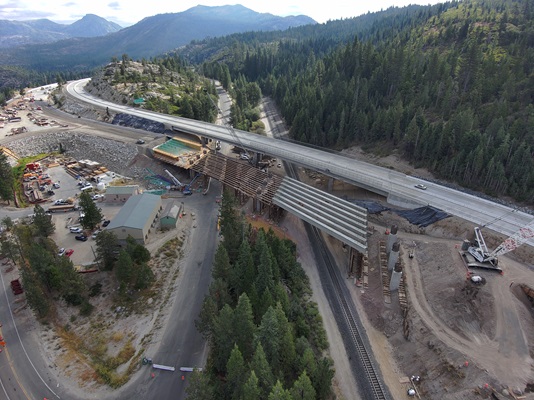 Aerial view of a highway bridge construction site in a mountainous, forested area. A completed elevated roadway curves across the scene while a second bridge is being built beneath it, with large concrete girders and wooden formwork in place. Construction equipment and materials are scattered around the site, including cranes, vehicles, and stacks of steel beams. A rail line runs through the lower portion of the image, and several small buildings sit near the construction zone. Pine-covered hills and distant mountains surround the area.