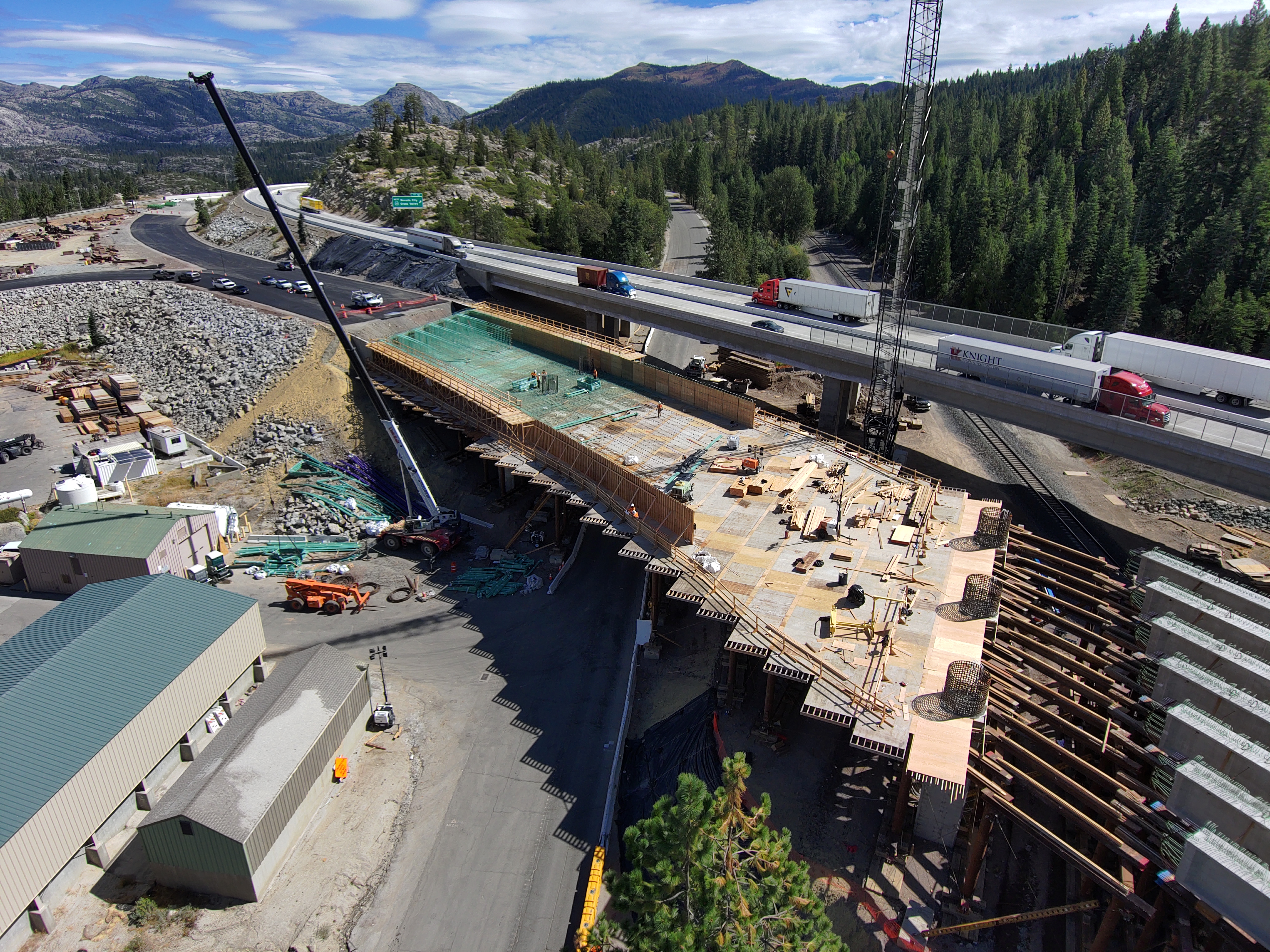 Aerial view of a large bridge construction site in a mountainous, forested area. Workers and equipment are positioned on a wide deck of wooden formwork and rebar, with cranes and machinery operating nearby. Semi‑trucks travel on the completed highway bridge above the active work zone. Construction materials are scattered around the site, and tall pine trees and rocky hills surround the area under a partly cloudy sky.