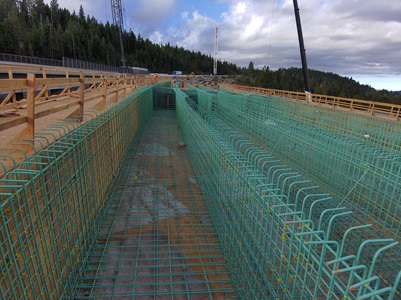 A large construction site shows long rows of green rebar cages arranged inside wooden formwork, ready for concrete placement. Cranes and equipment are visible in the background, with workers further down the structure. The area is surrounded by dense evergreen forest under a cloudy sky.