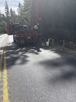 A roadside construction crew works beside a large truck equipped with a tall post‑driver. Workers in high‑visibility safety gear install new metal guardrail posts along the forested edge of a two‑lane road. Sunlight filters through the trees, casting shadows across the pavement.