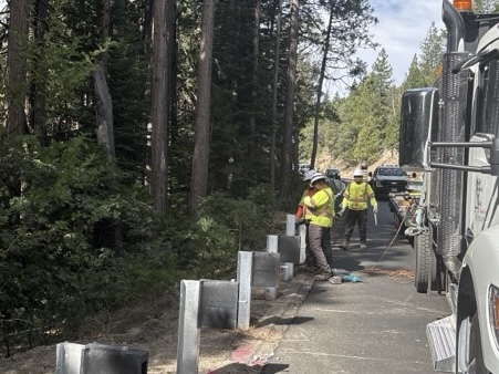 Construction workers in high‑visibility safety gear stand along a roadside installing new metal guardrail posts. A large work truck is parked nearby, and the scene is bordered by tall trees and forest vegetation under partly cloudy skies.