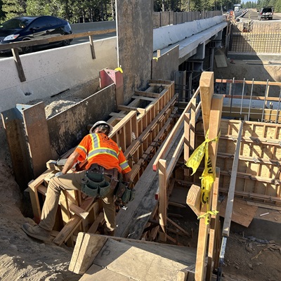 A construction worker wearing a hard hat and bright orange safety vest builds wooden formwork beside a bridge structure. The worker is positioned on a narrow ledge, adjusting the lumber framing around rebar and concrete components. Vehicles pass on the roadway above, and yellow caution tape is tied to parts of the formwork. The scene is set in a forested area with sunlight filtering through the trees.