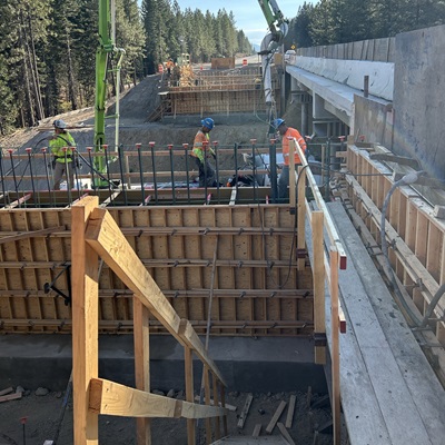 Construction workers wearing safety gear stand on wooden formwork beside a bridge structure, guiding a concrete pump hose as rebar is set in place. The scene shows multiple levels of timber framing, exposed rebar, and equipment along an active roadway. Tall pine trees line the background under clear daylight.
