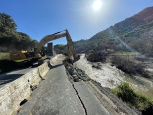 Photograph showing the slip out on State Route 84 near Sunol. There is a creek on the right side of the damaged road. There is a barrier separating the roadway from the damaged portion. There is an excavator on the left side that is reaching over the barrier to work on the damaged portion of the roadway.