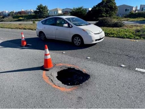 Photograph of a sinkhole in the far-right lane of State Route 35 in San Mateo County. There are orange traffic cones closing the lane around the hole while a white sedan passes in the left-hand lane.
