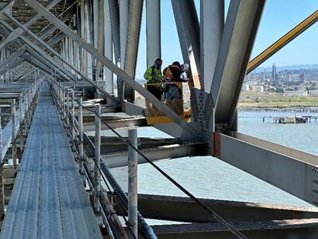 Two Caltrans workers perform an inspection of the steel beams of a bridge below the road deck.