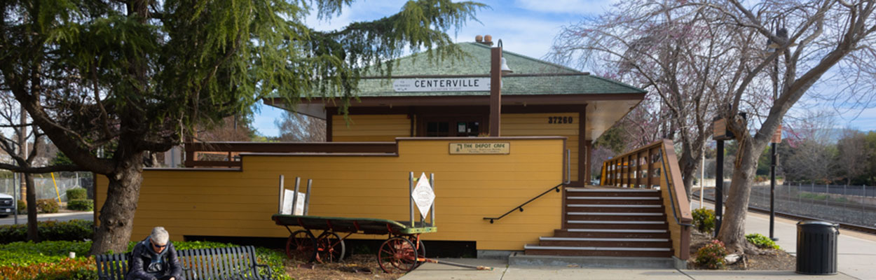 Banner image of the Fremont -Centerville Amtrak station building with a passenger sitting on a park bench.