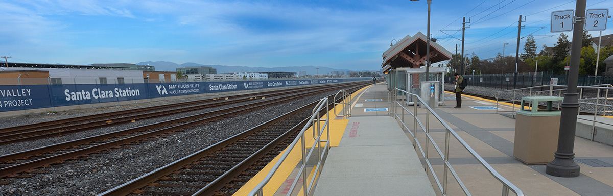The station platform at Santa Clara University Station with two sets of train tracks running along the left side of the image. On the opposite side of the tracks from the platform is a fence featuring a banner that reads Santa Clara Station.