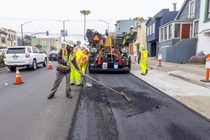 A Caltrans crew uses heavy equipment and hand tools to apply new asphalt on southbound 19th Avenue (State Route 1) on May 10, 2026. The left lane remains open to vehicle traffic.