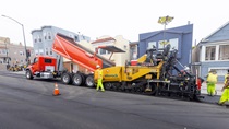 A truck dumps asphalt into the front of heavy equipment used to apply new asphalt on southbound 19th Avenue (State Route 1) on May 10, 2026.