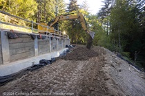 An excavator parked on the roadway dumps a bucket of soil into the work area downslope of the roadway.