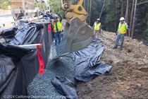 An excavator pours a bucket load of gravel onto a tarp next to the roadway while three workers wait with shovels.
