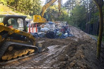 Two Caltrans workers distribute gravel around the worksite with shovels while an excavator and a front loader are operated nearby.