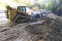 A front loader pushes soil in the work area near the repaired roadway on State Route 84 in San Mateo County while a group of Caltrans employee use shovels to distribute gravel.