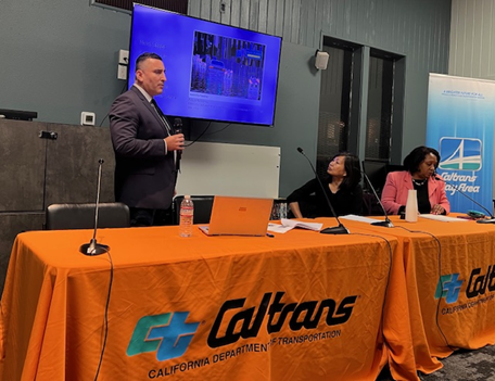 Caltrans representatives at the US-101, State Route 1 (SR-1) Sea-Level Rise project in Marin City on Thursday, Feb. 29. One representative is standing behind a table with an orange Caltrans tablecloth and talking to the audience about the presentation on-screen behind him. Two additional representatives sit behind another table with an orange Caltrans tablecloths.