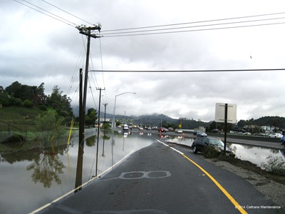 Photograph of flooding at the exit for Donahue Street on southbound US 101 near Marin City. Portions of the offramp are covered in water and a car stopped in the dirt separating the offramp from the highway.