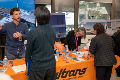 Caltrans representatives speaking with attendees at the US-101, State Route 1 (SR-1) Sea-Level Rise project in Marin City on Thursday, Feb. 29. Two representatives stand behind a table with an orange Caltrans tablecloth and talk with two attendees standing on the other side of the table about the proposed project.