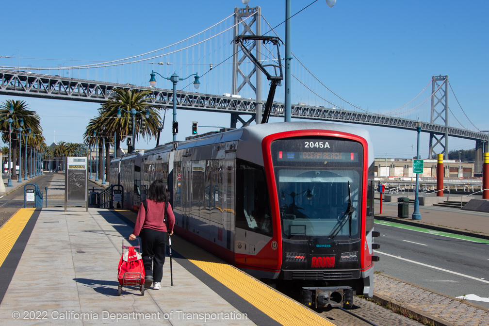 Person with a roller luggage walking towards a Muni Metro Light Rail train.