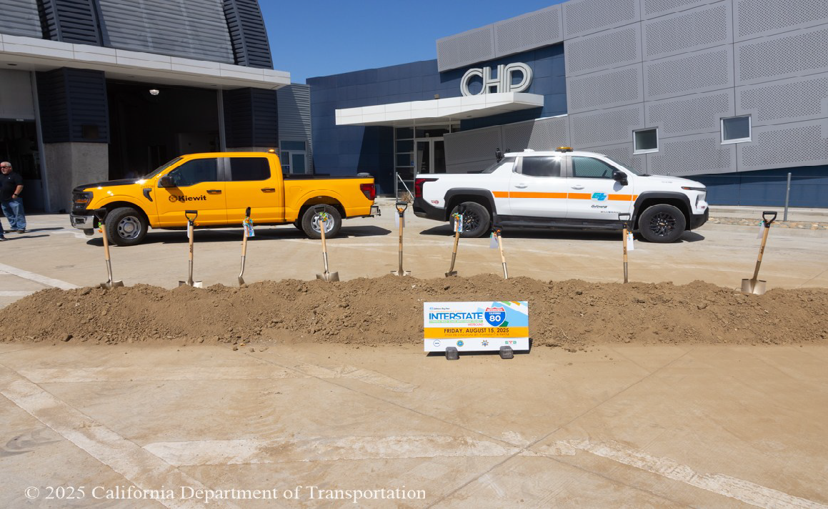A sign for the Interstate 80 truck scales project groundbreaking ceremony sits in front of a long pile of dirt with nine shovels stuck in it. Behind the pile of dirt, a yellow pickup truck and a white Caltrans pickup truck are parked in front of a CHP building.