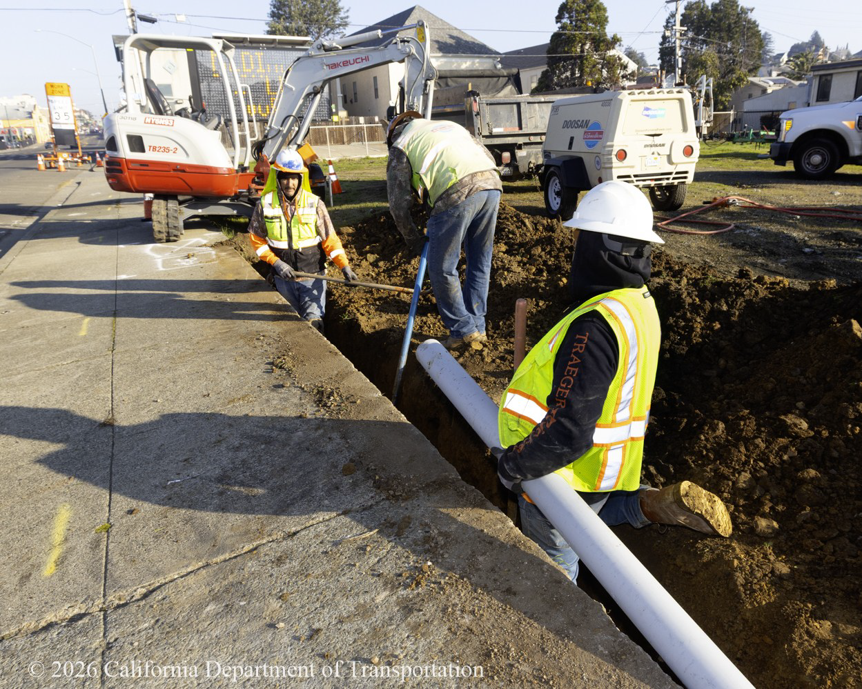 Three Caltrans crew members install a pipe in a trench along the sidewalk on State Route 29.