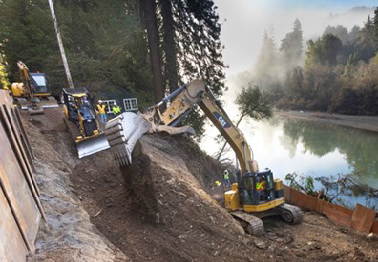 An excavator builds an embankment between a temporary retaining wall and the Russian River on Route 116 between Duncans Mills and Monte Rio.