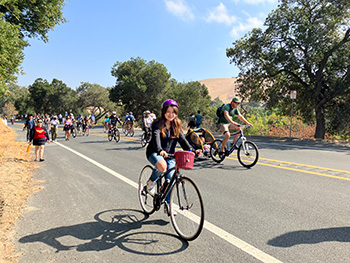 Photograph of a group of bicyclists and pedestrians traveling on a tree-lined road.