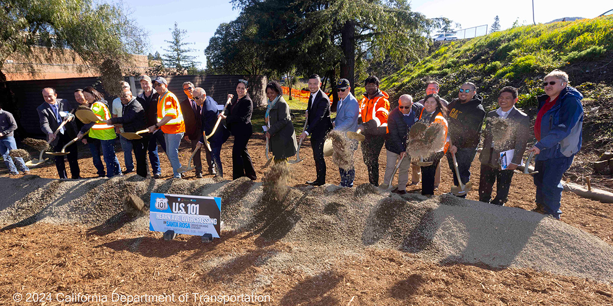 A group of representatives from Caltrans, Sonoma County Transportation Authority, and local elected officials stand behind a pile of gravel and toss shovelfuls of dirt as part of the US 101 Hearn Ave Overcrossing Project's groundbreaking ceremony.